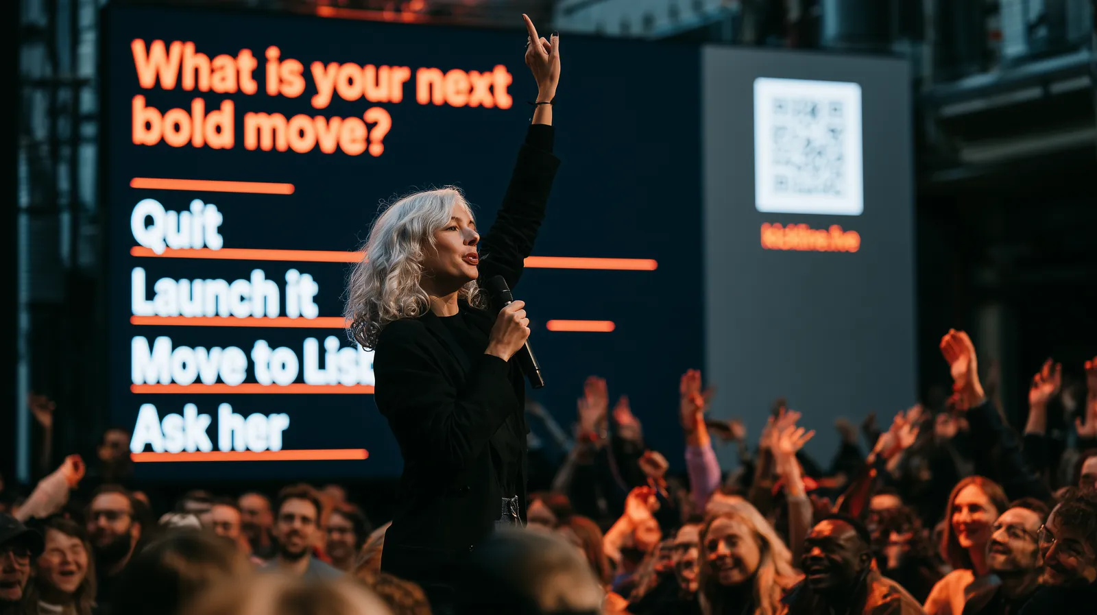 A keynote speaker raises her hand and the audience raises theirs, the answers on a screen behind her